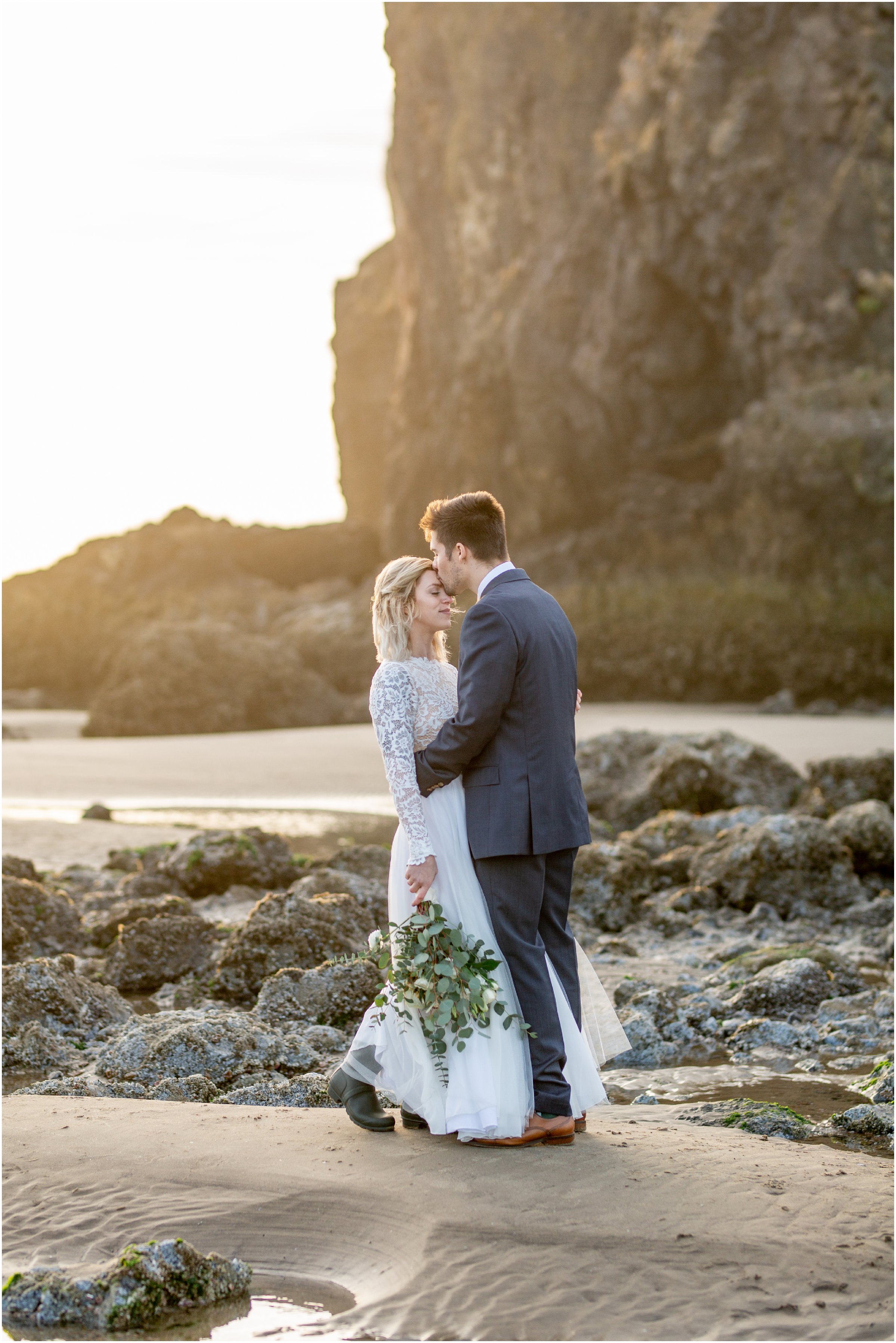groom kisses bride on the forehead on the beach as sun shines through the rocks during sunet by cannon beach elopement photographer