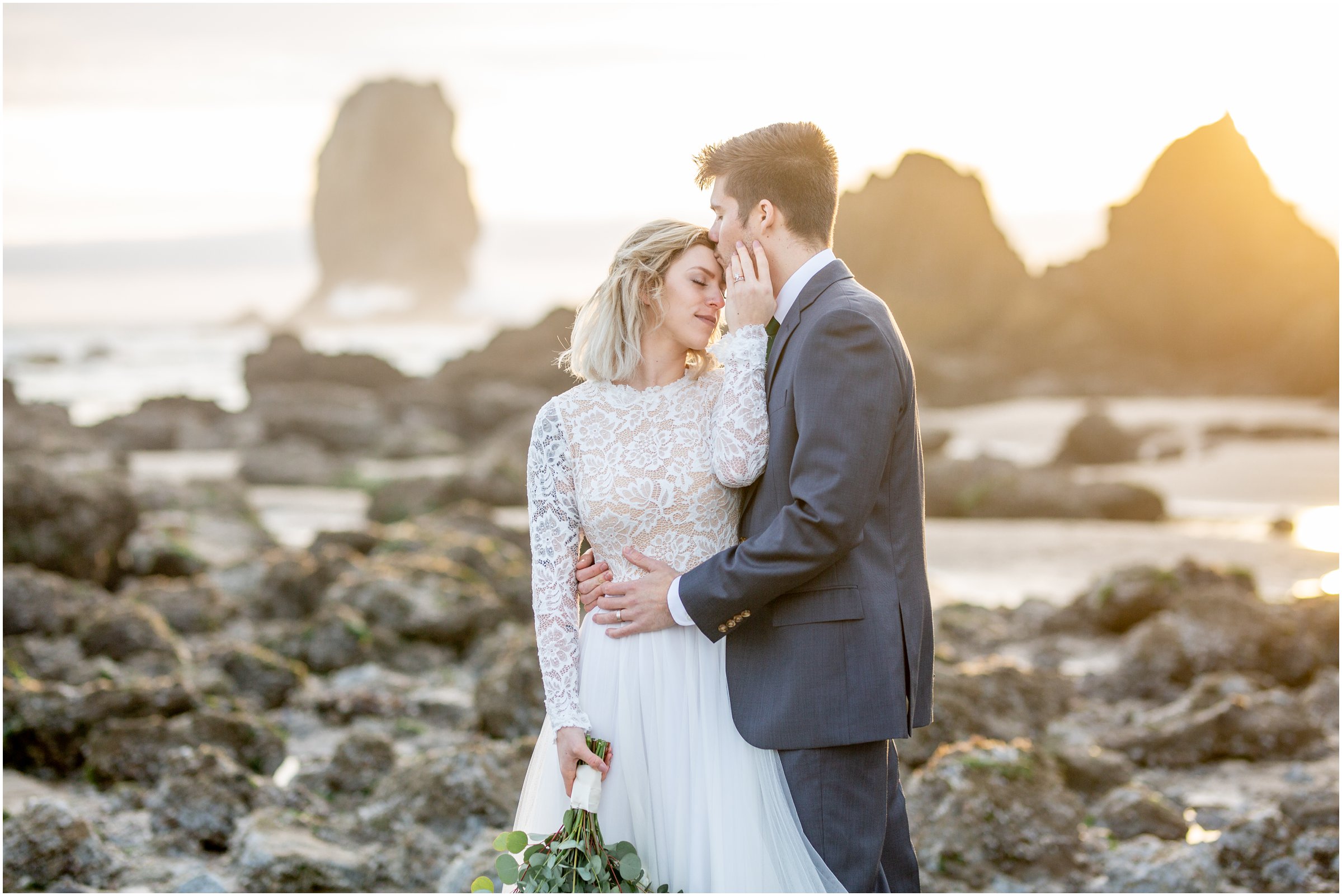 bride and groom snuggle on the beach as the sun shines through the rocks during sunset