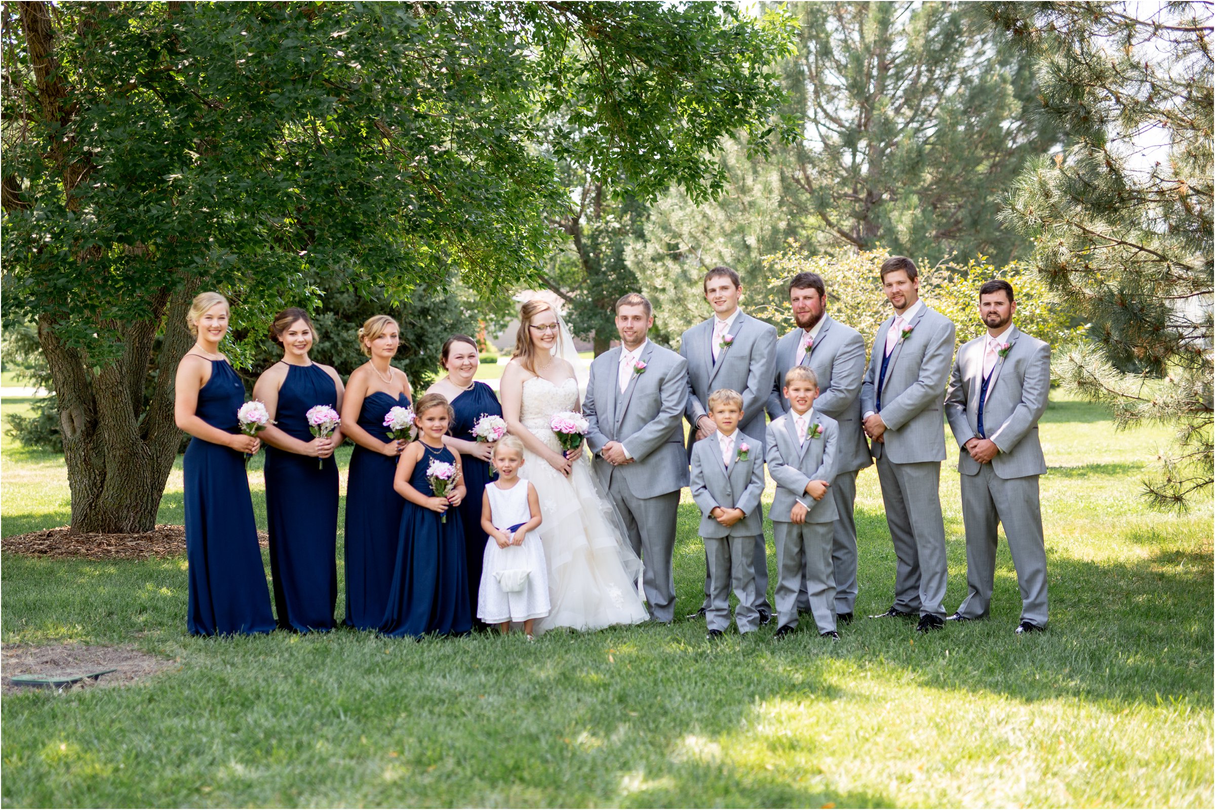 bride and groom pose with her bridesmaids in navy blue dresses with pink flowers and groomsen in gray before their wedding at holdrege trinity church