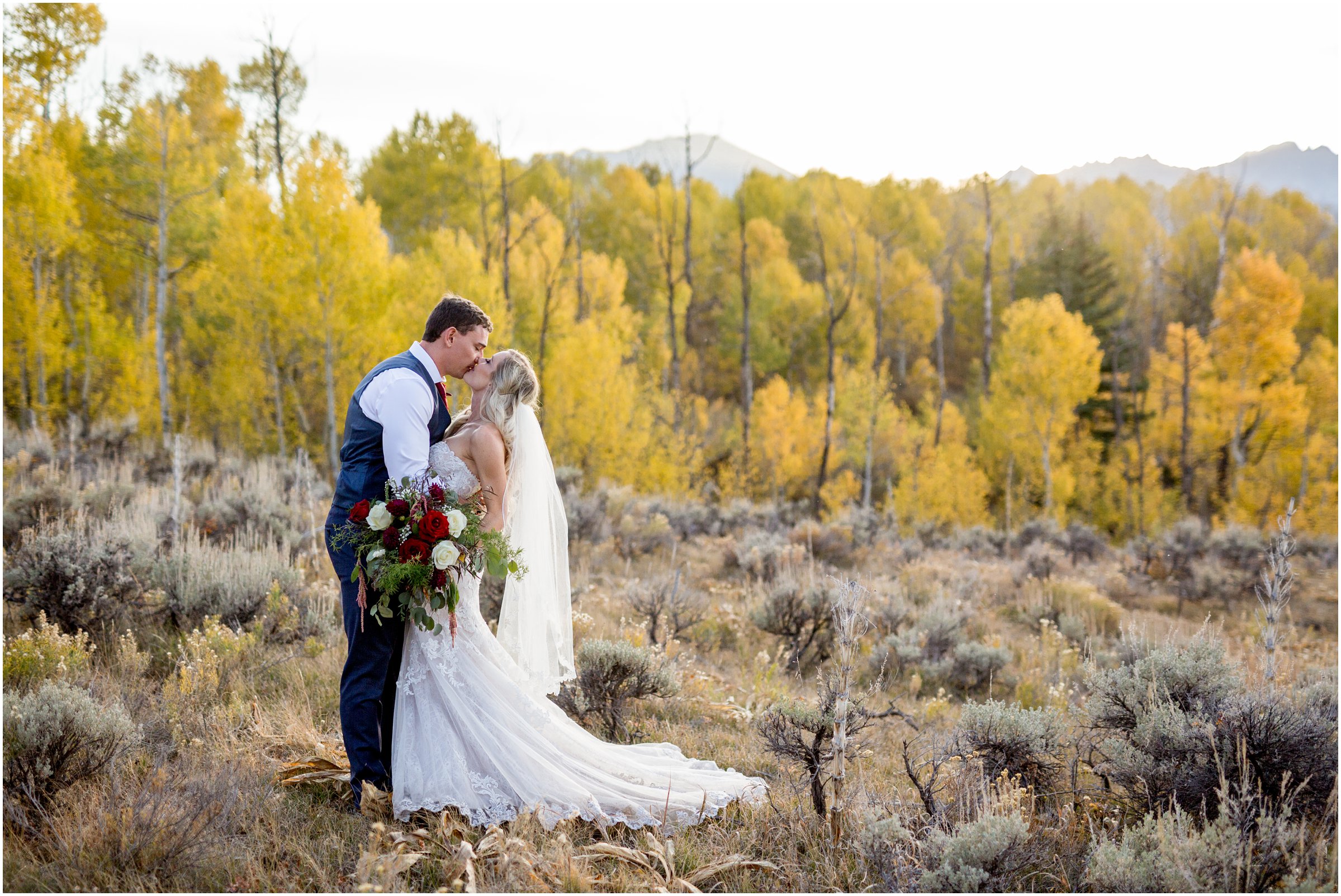 bride and groom kiss in an open field with fall colored trees and mountain peaks in the background