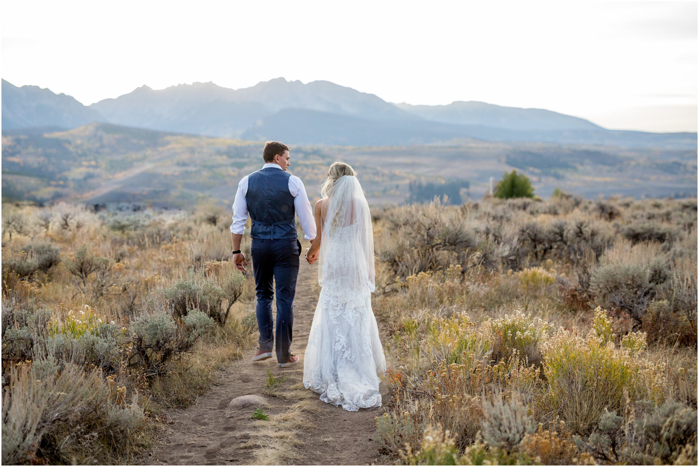 bride and groom walk down a path together toward a mountain range holding hands