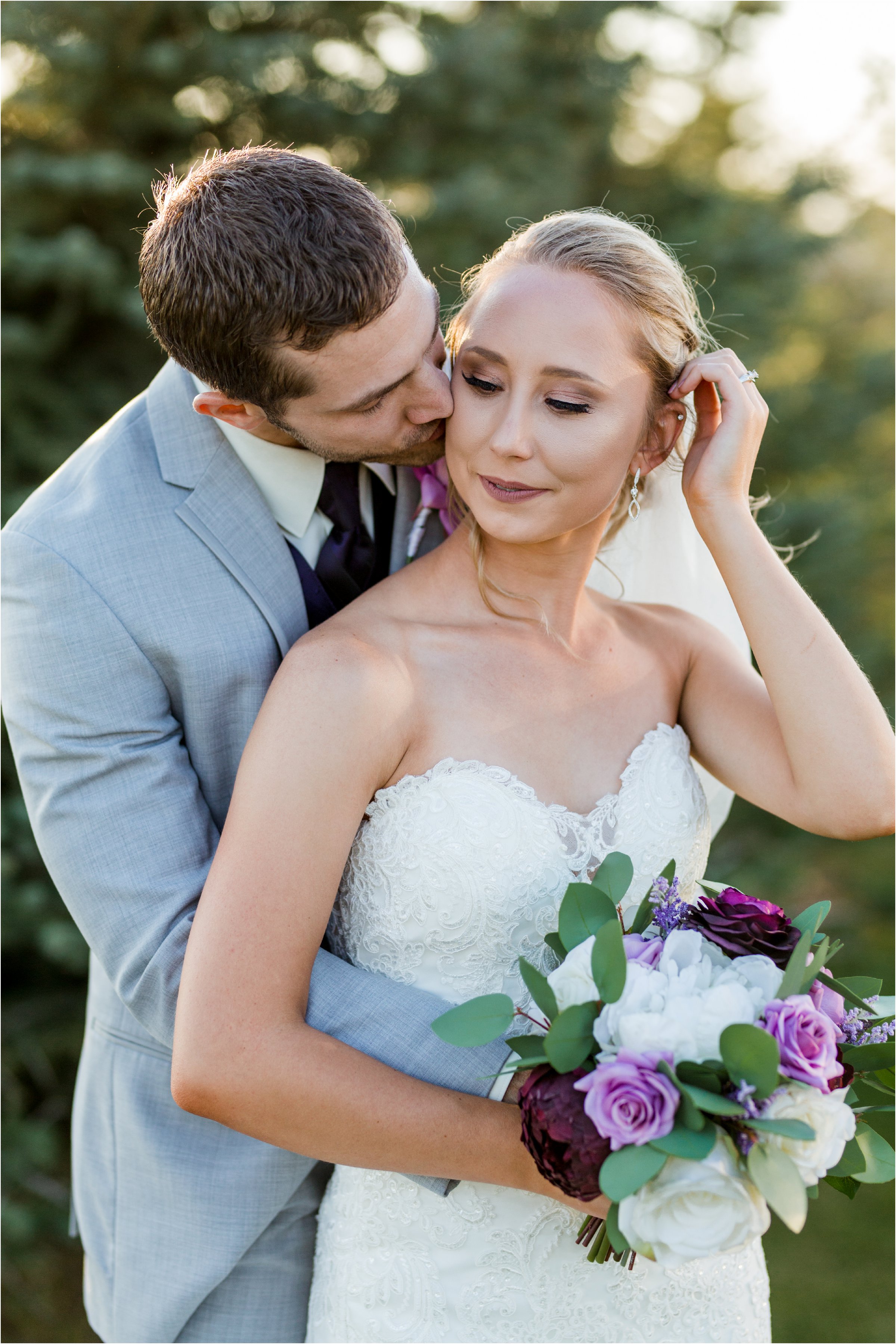 groom kisses bride on the cheek in front of trees as the golden light of sunset shines through
