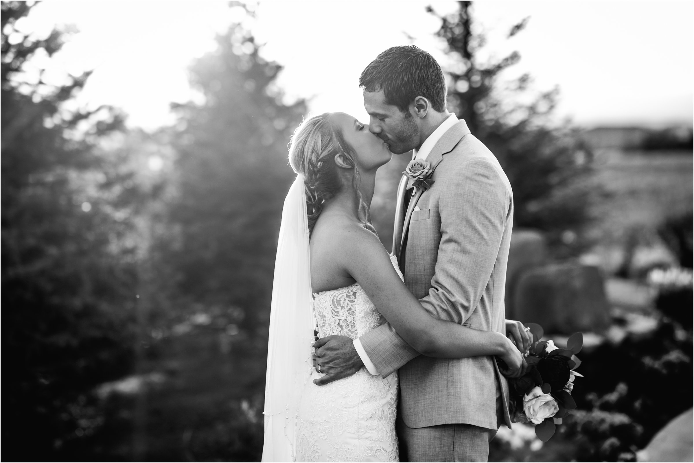 black and white photo of bride and groom snuggle in front of trees as the golden light of sunset shines through