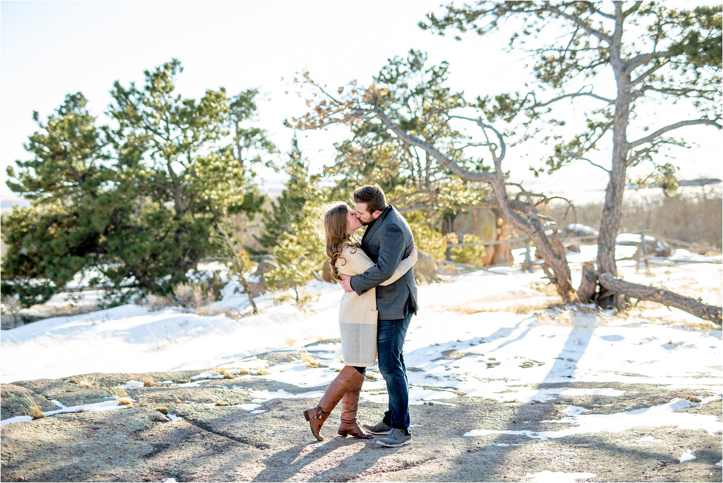 bride and groom kissing on a large rock with trees and snow in the background for the engagement session
