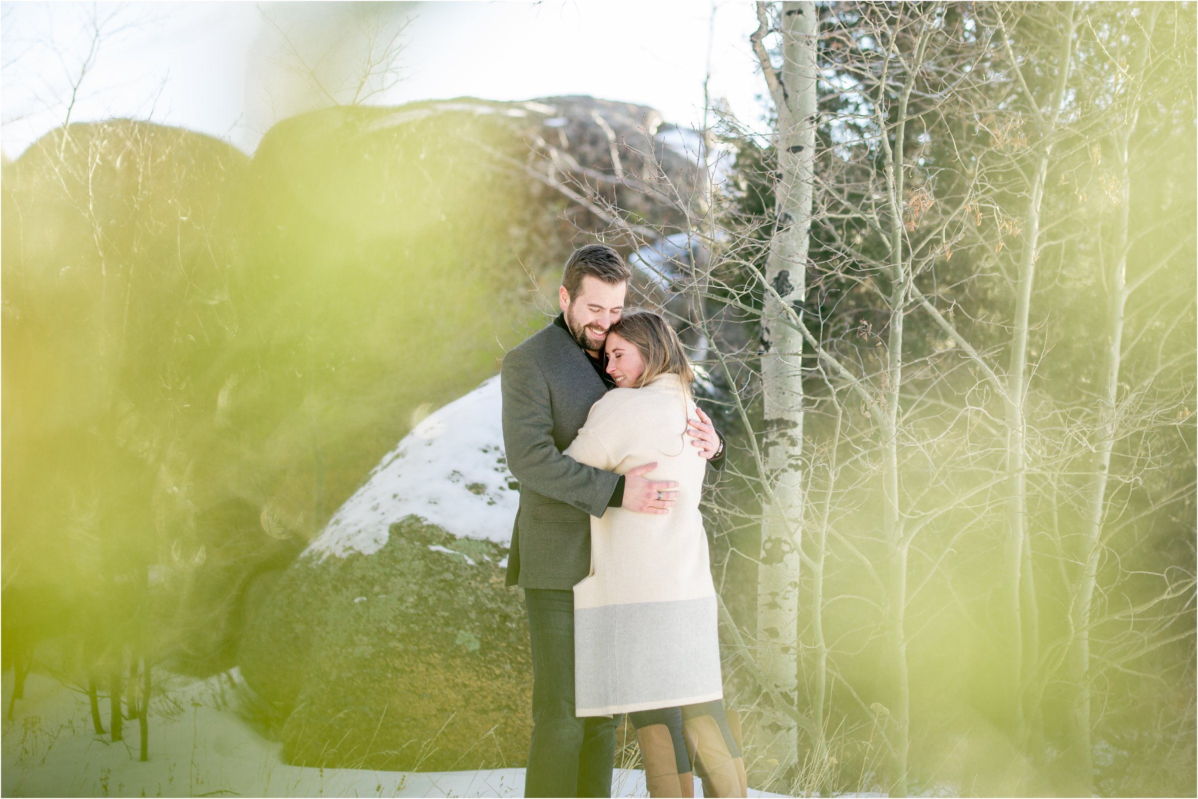 engaged couple with out of focus greenery in the foreground cuddling with rocks and trees in the background