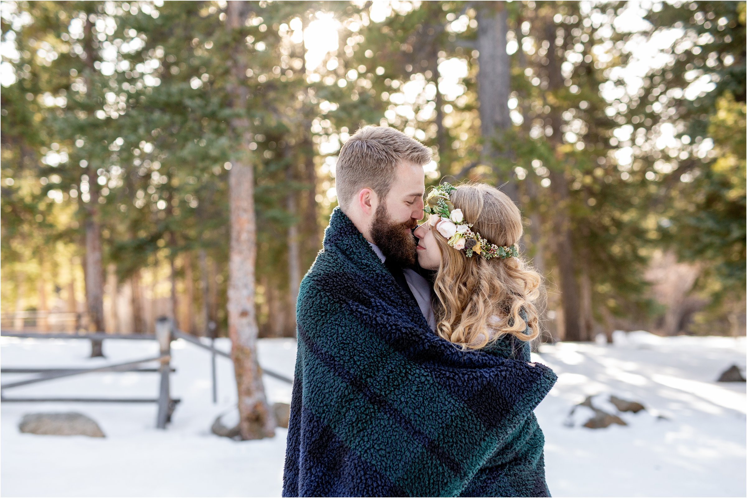 Bride and groom snuggling faces wrapped in blanket in front of evergreen trees