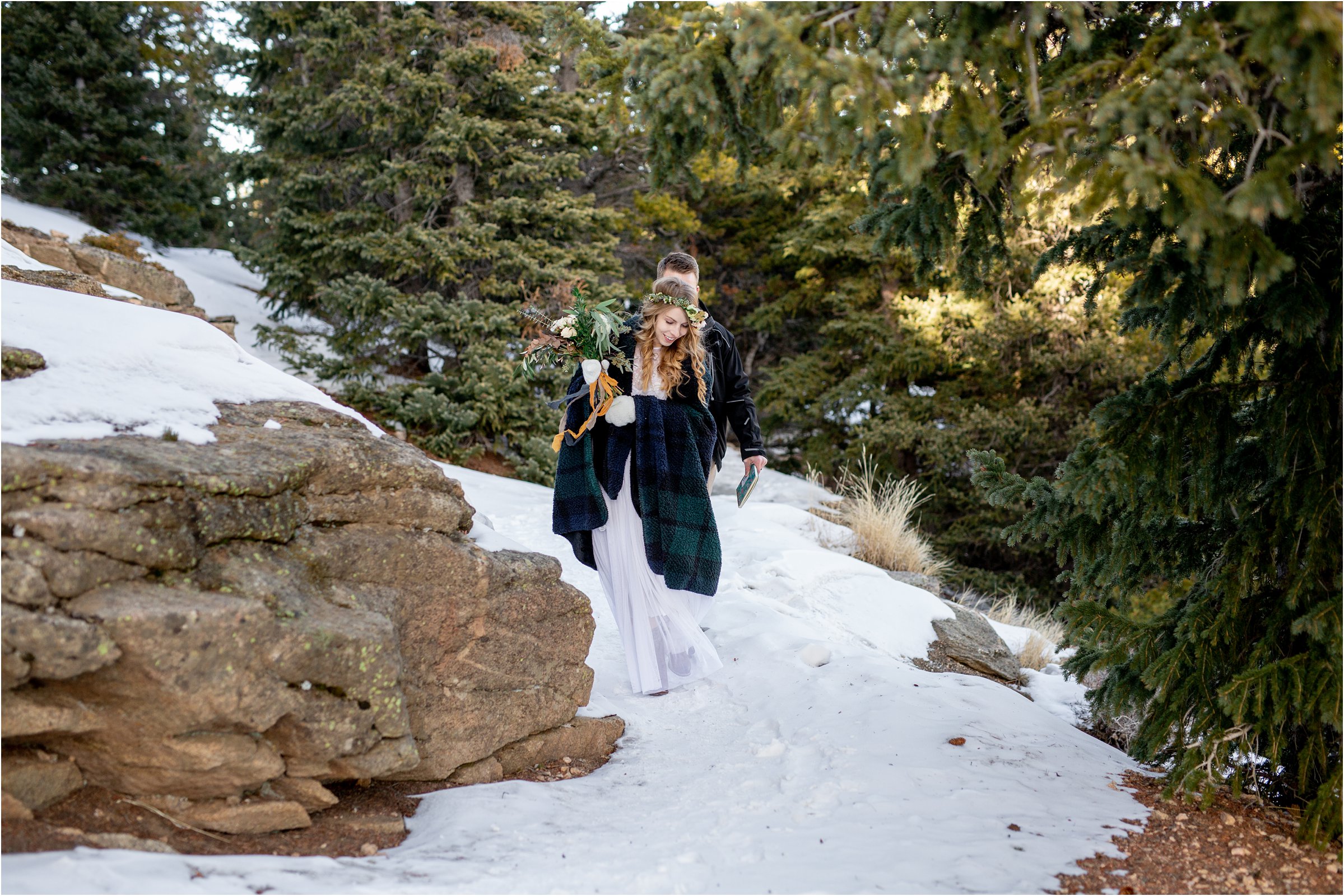 Bride and groom walking through trees on snowy path in warm clothes and blanket