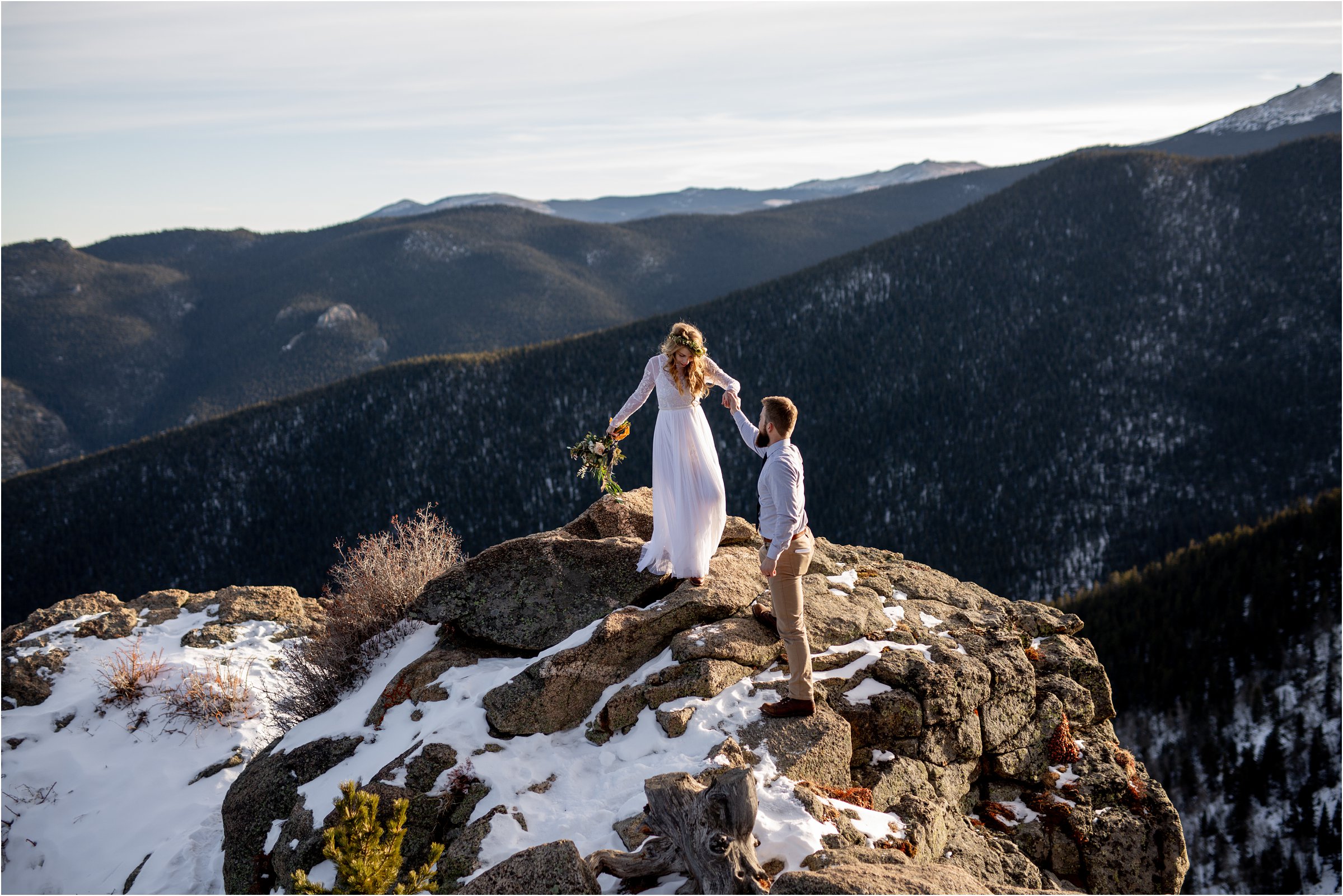 Bride in wedding dress with bouquet being helped down off mountain by grooom