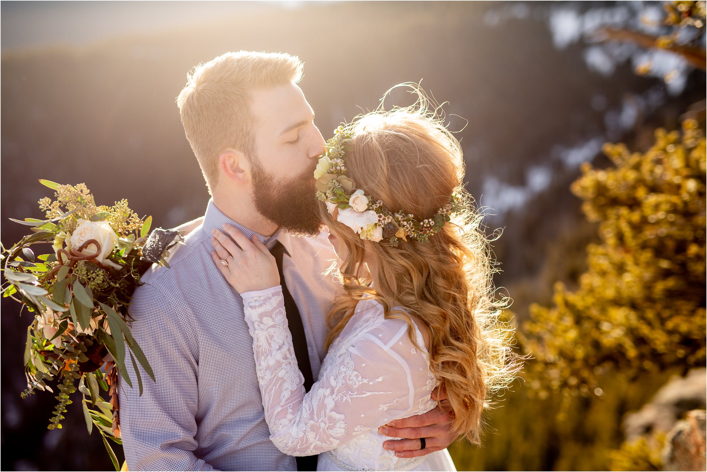 Bride and groom on mountain with large bouquet while groom kisses bride's forehead