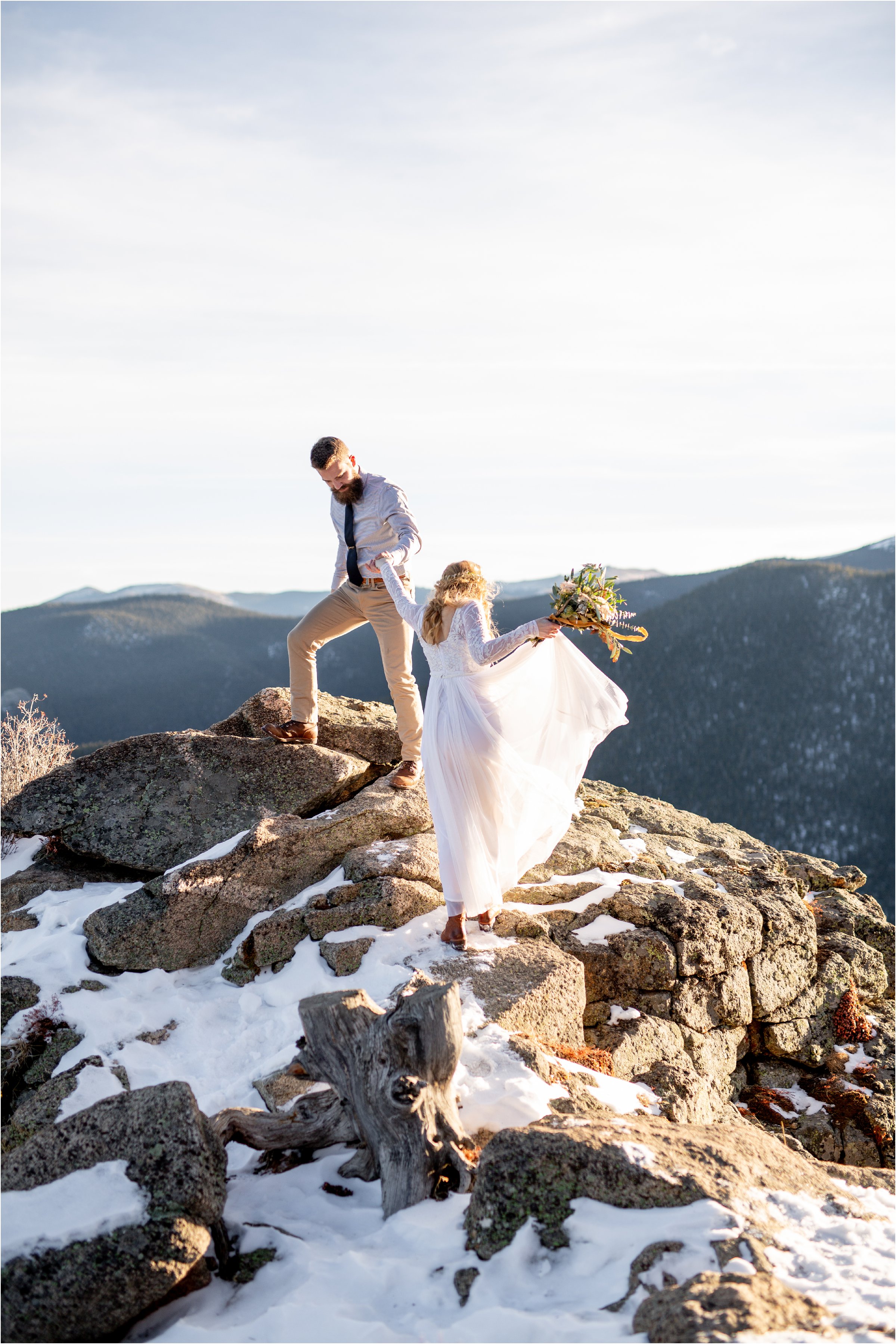 Groom helping bride onto mountain top with large bouquet for their Denver Elopement