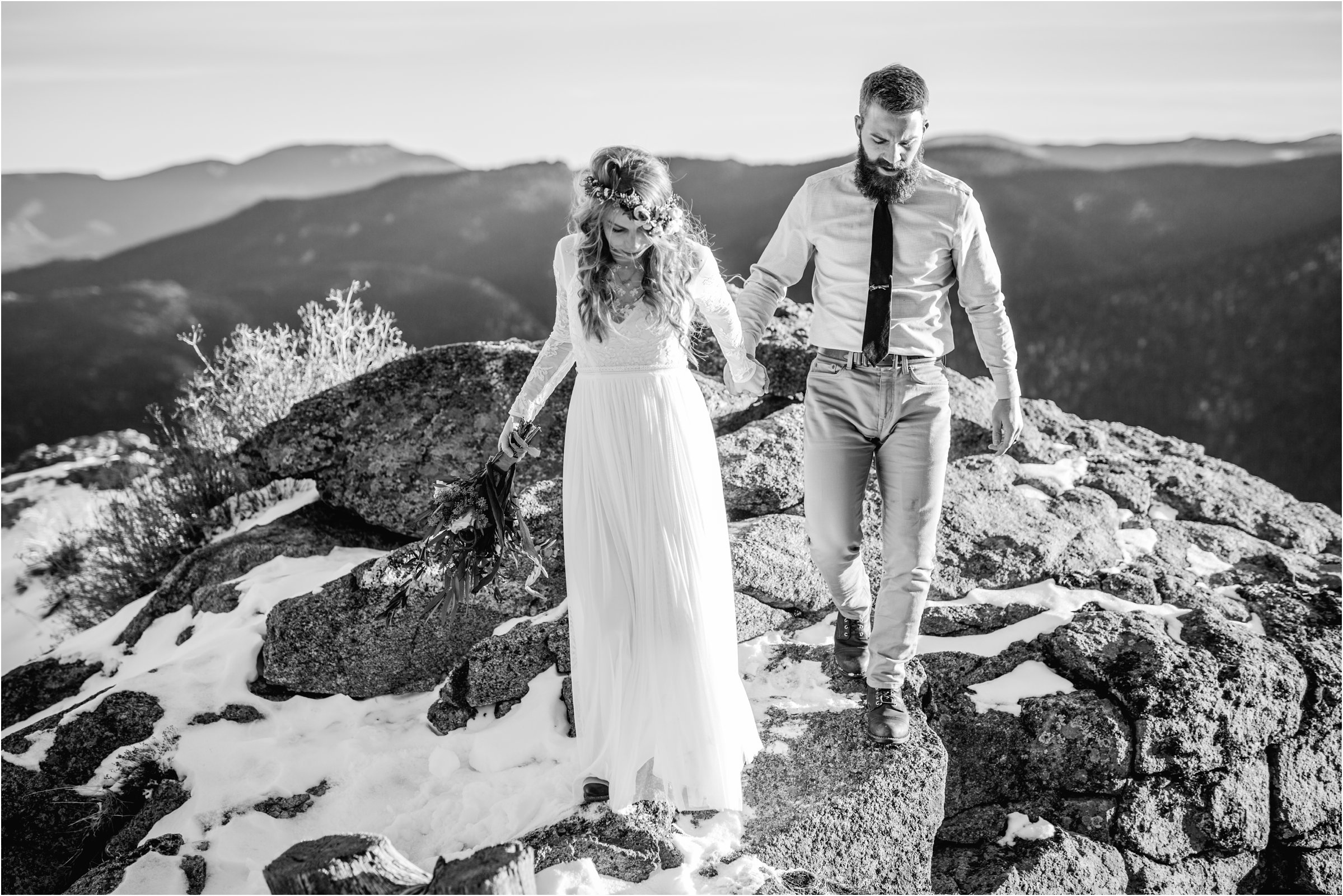 Groom and bride in wedding dresss walking together off a mountain top with large bouquet and flower crown