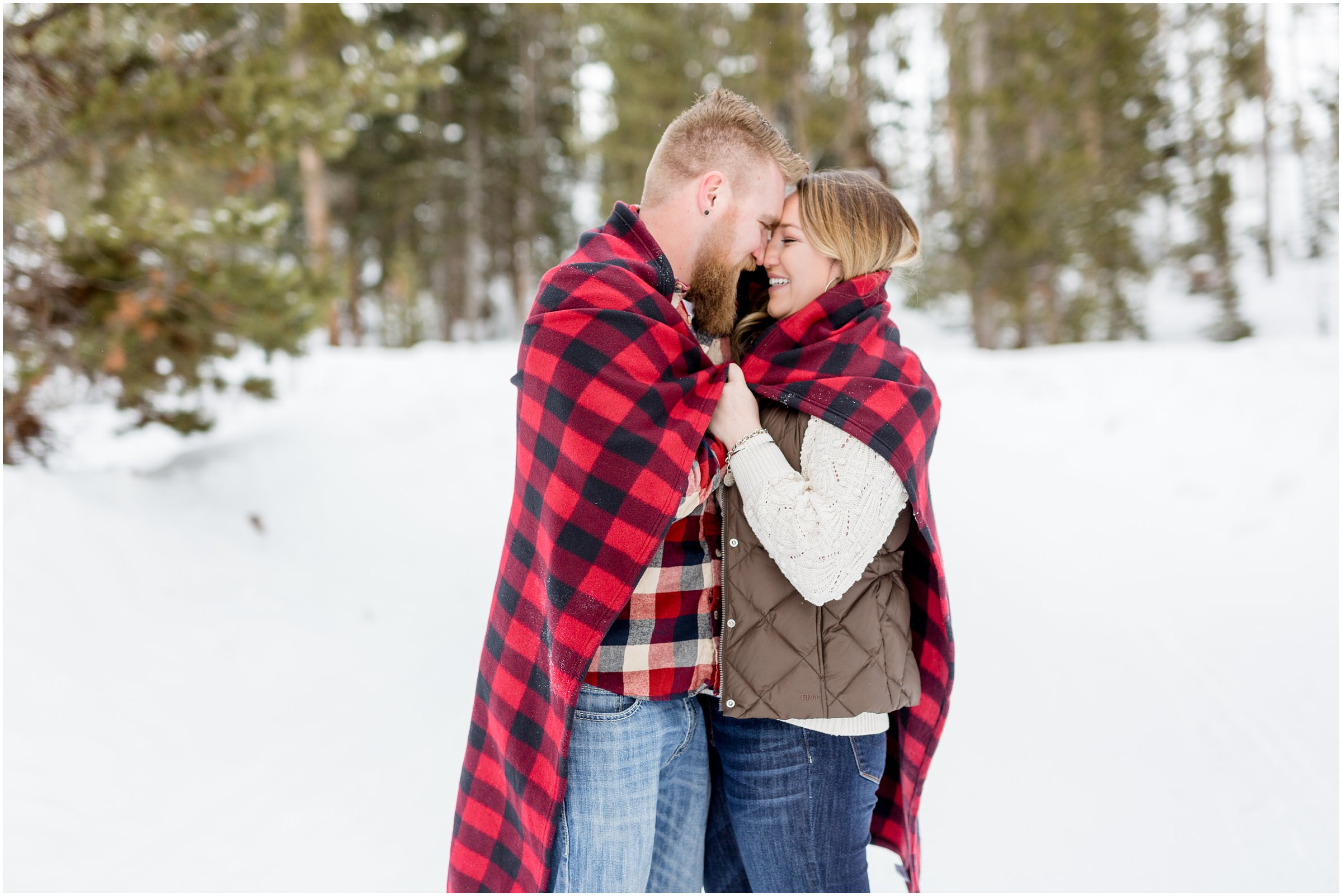 Man and woman in winter clothing pose as bride and groom snuggle in a red plaid blanket 