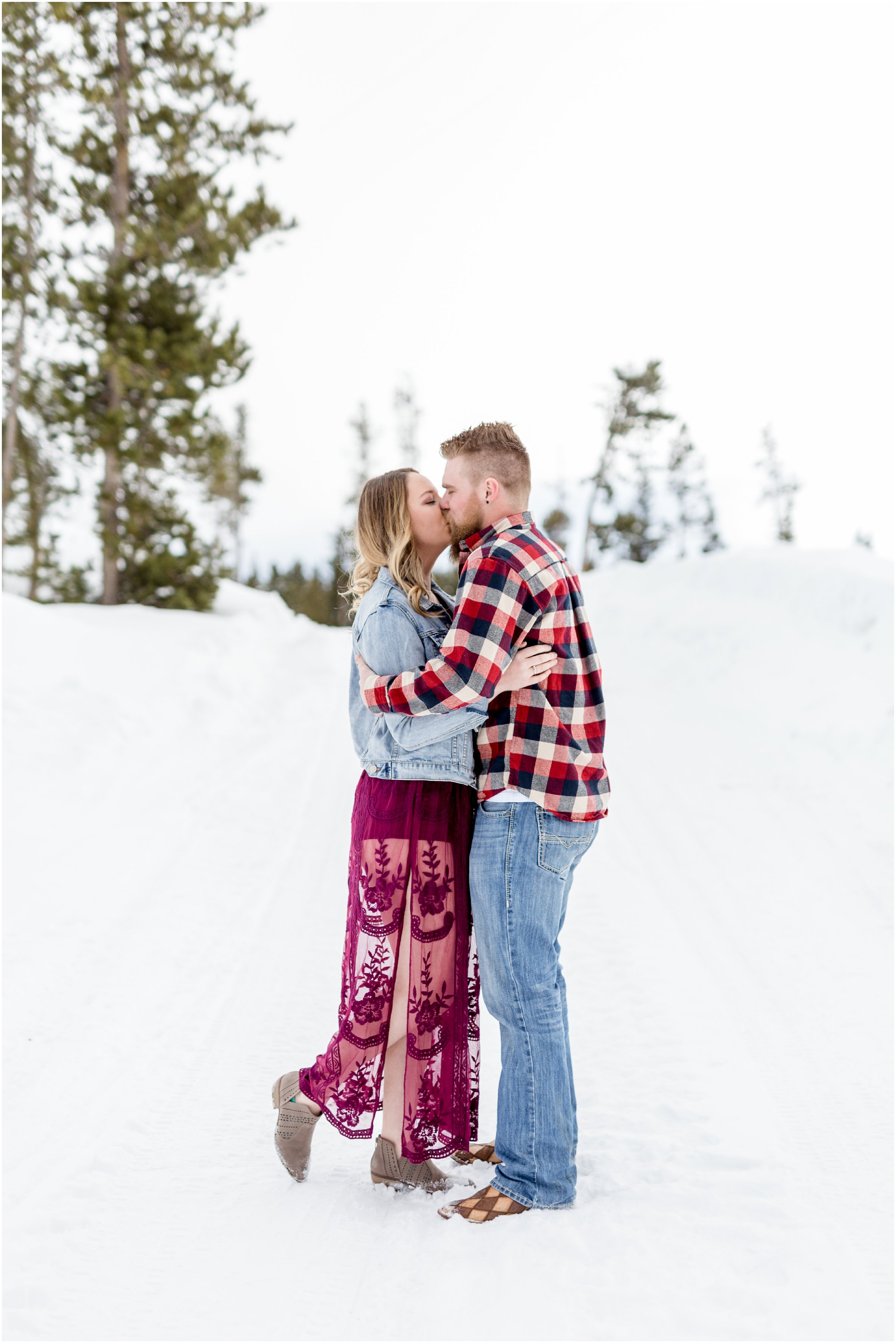 man and woman kiss in front of a snow drift and trees