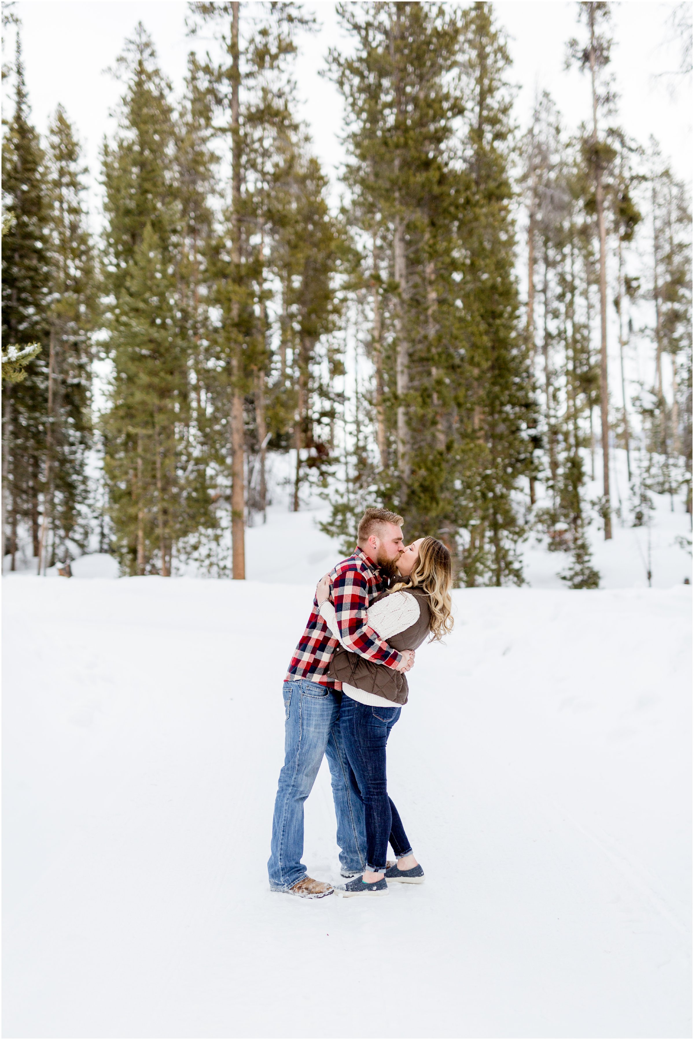 groom kisses bride on the lips in front of snow and green trees