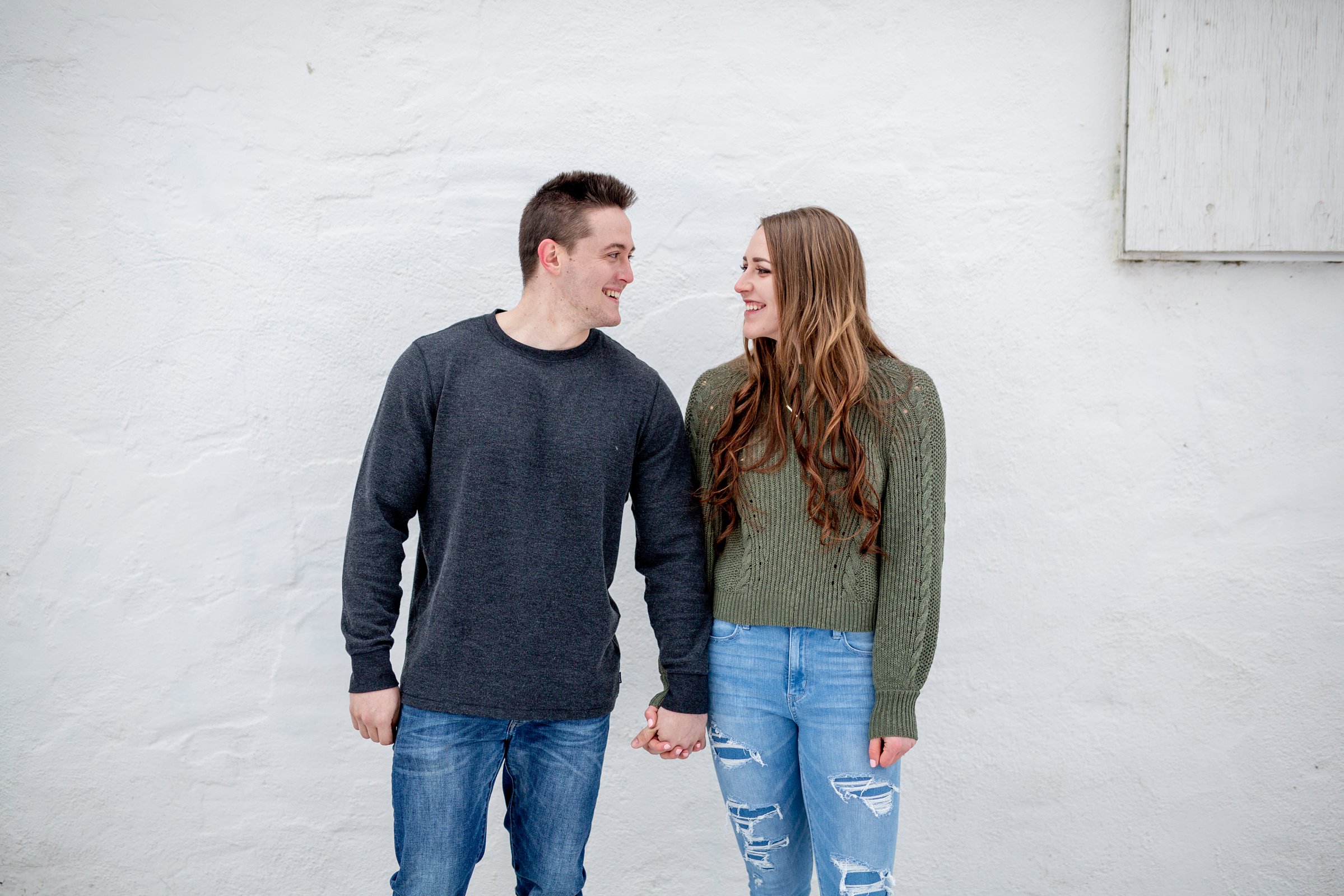 engaged man and woman in front of white wall holding hands and looking at each other for fort collins engagement session