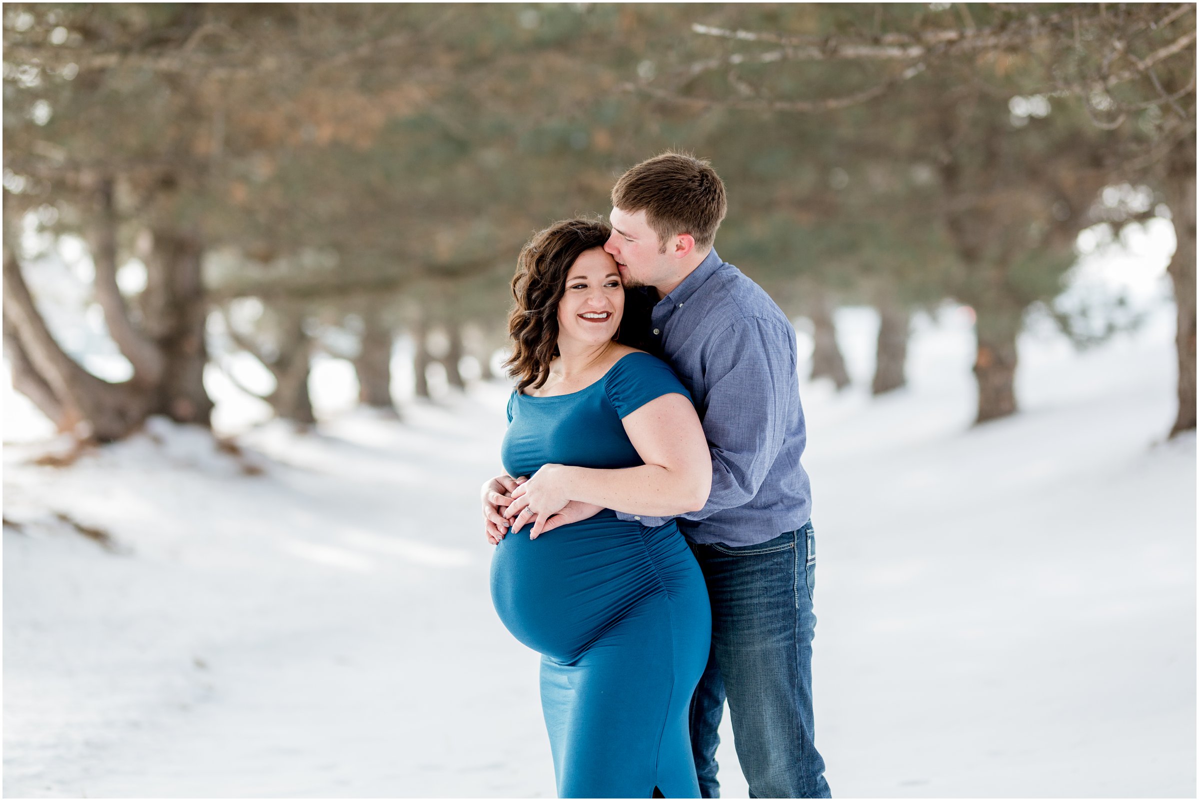 pregnant woman in a blue dress being kissed by her husband in a row of trees