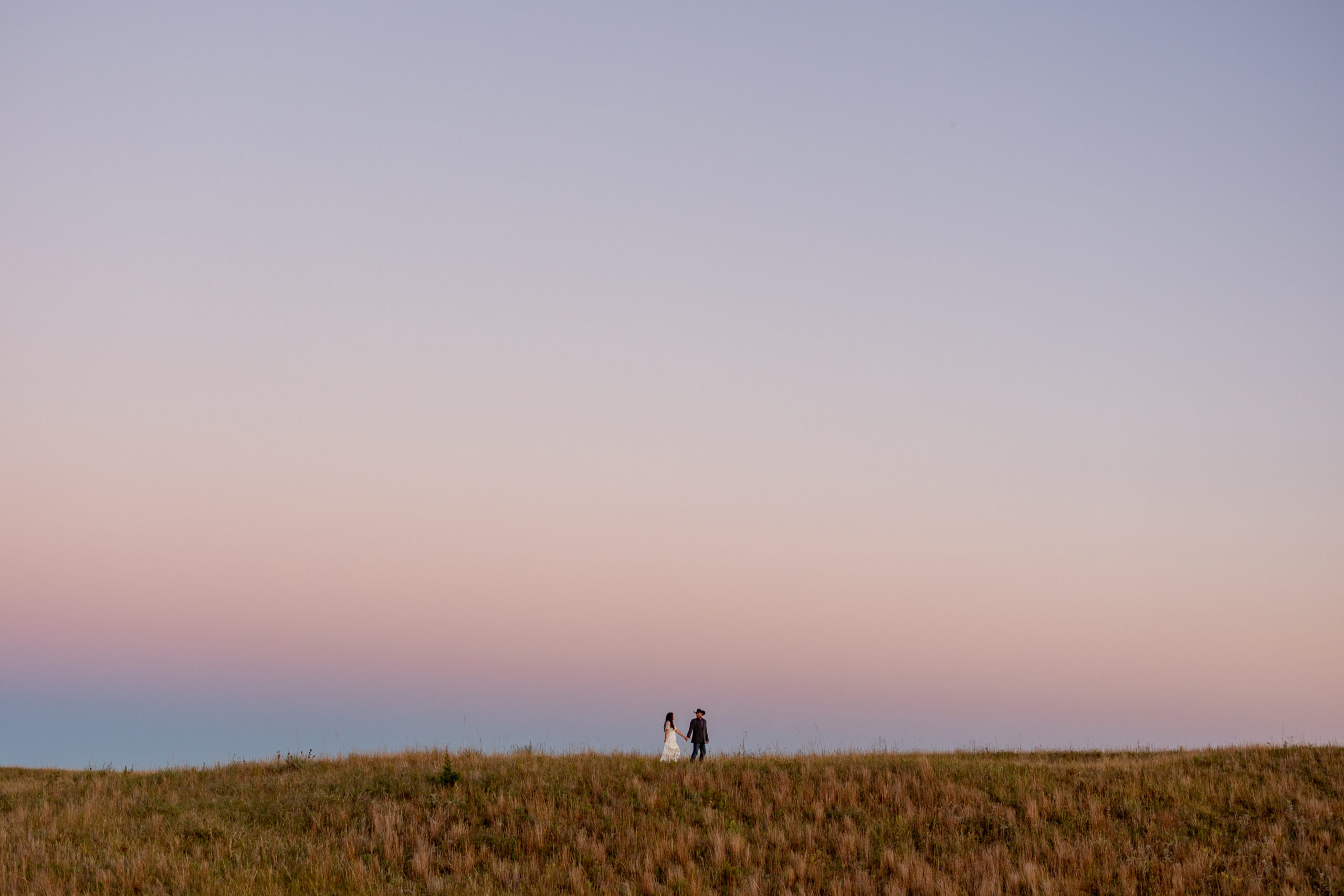 couple against a cotton candy pink and blue sunset sky walking along a ridge in the nebraska sandhillls during their engagement session